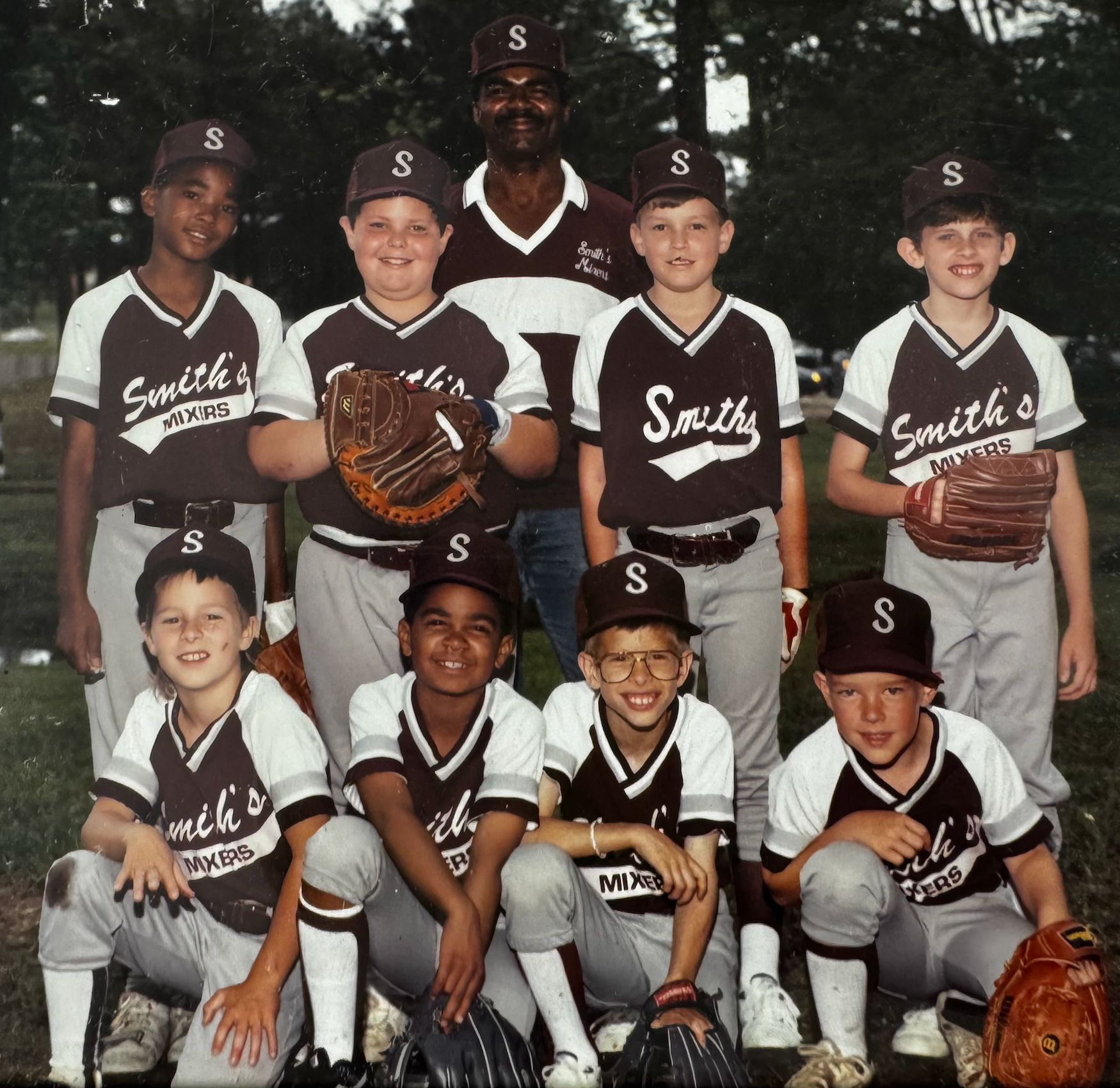 Youth baseball team, wearing brown/white uniforms, pose with coach in front of trees.