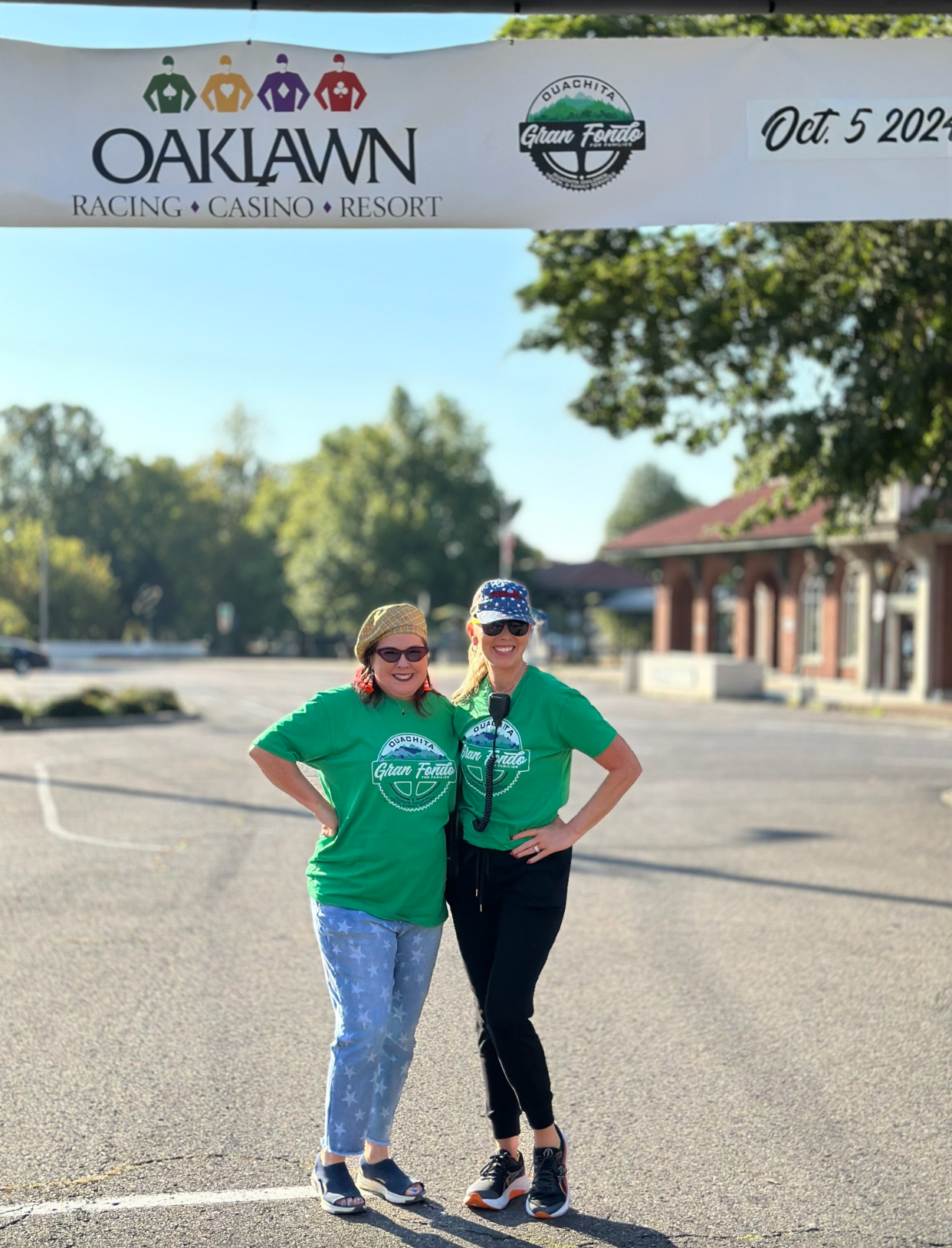 Two women pose under a race banner at Oaklawn Racing Casino Resort. Green shirts, blue sky.