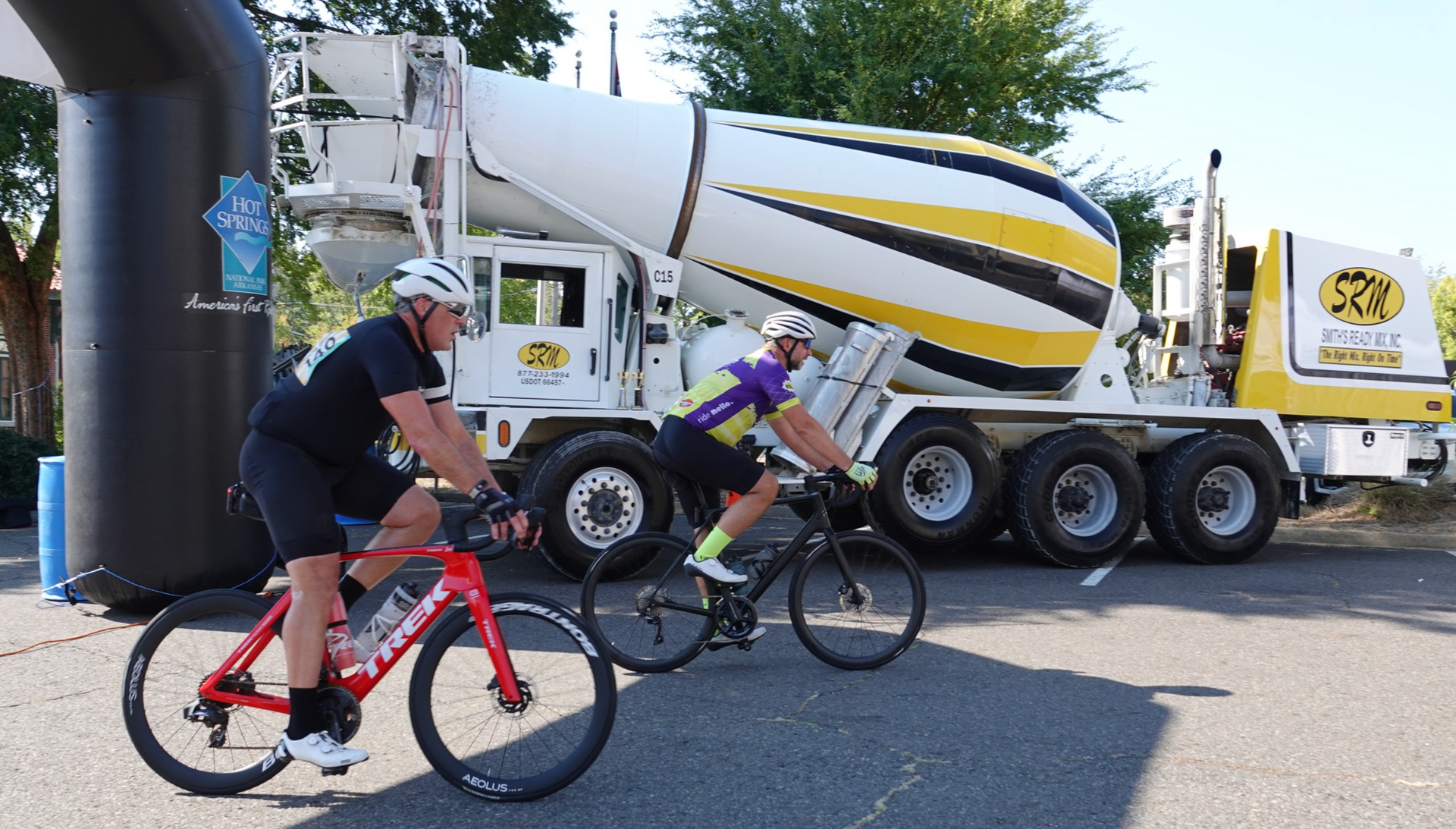Two cyclists on road bikes pass a large cement truck at an outdoor event, under a starting arch.