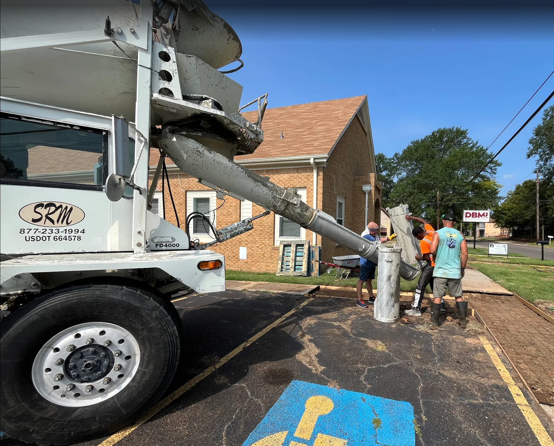 Concrete truck pouring concrete into a parking area near a brick building. People are working.