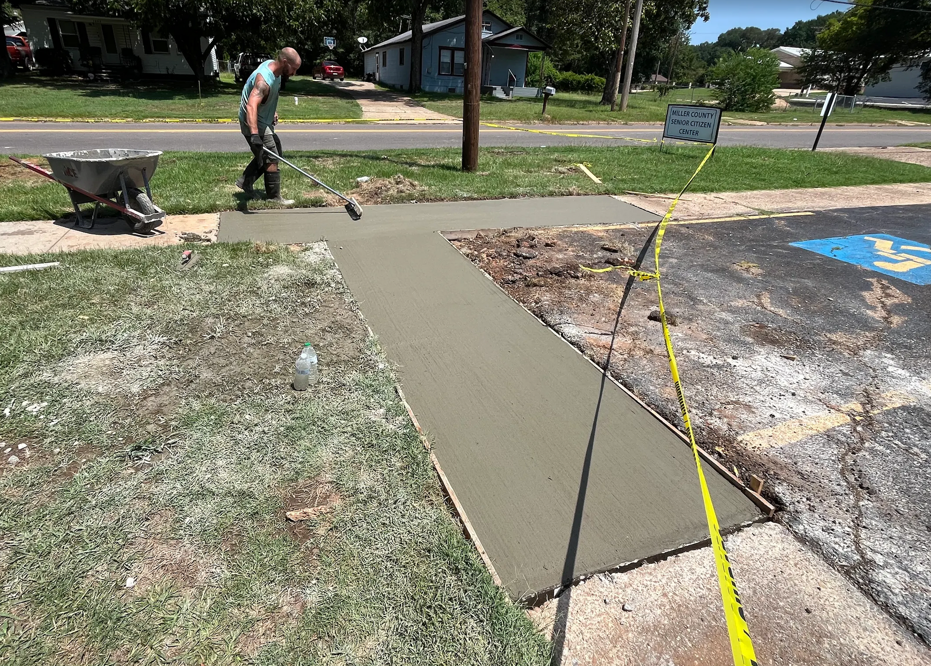 Man smoothing wet concrete sidewalk. Wheelbarrow, tape, and building in background.