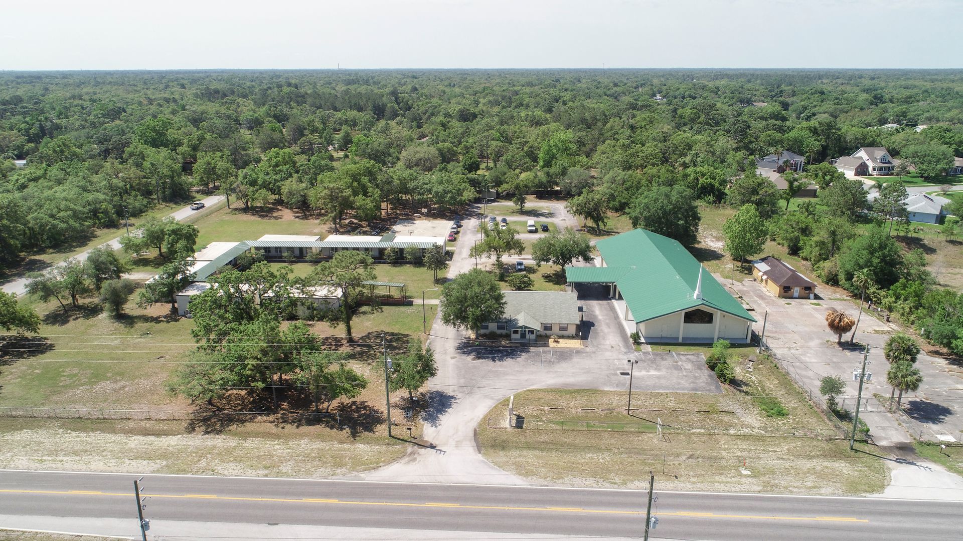 Aerial view: Green-roofed building and long white structure surrounded by trees, with a road in front.