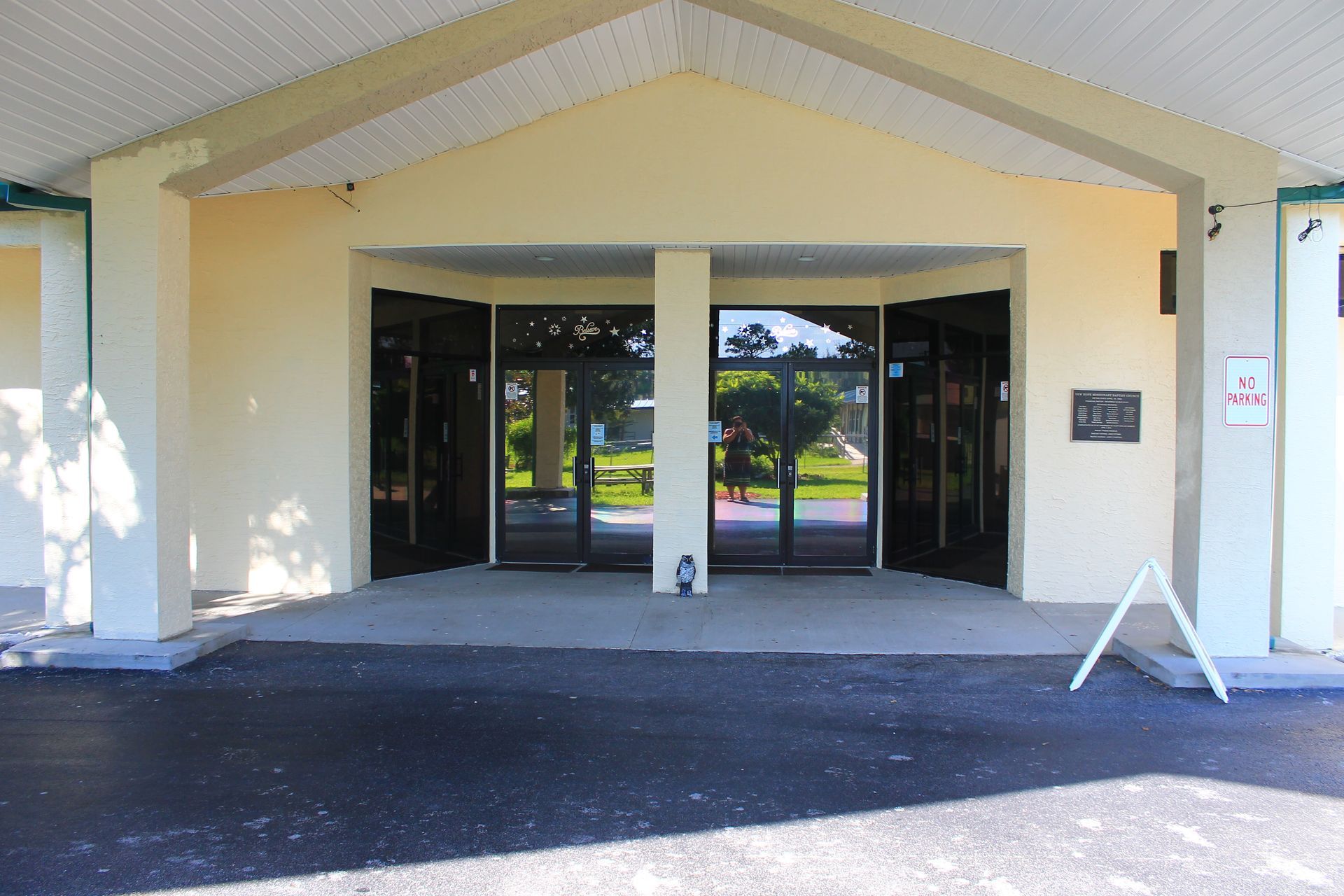 Exterior of a building with glass doors, tan walls, and a concrete entrance. An A-frame sign sits to the side.