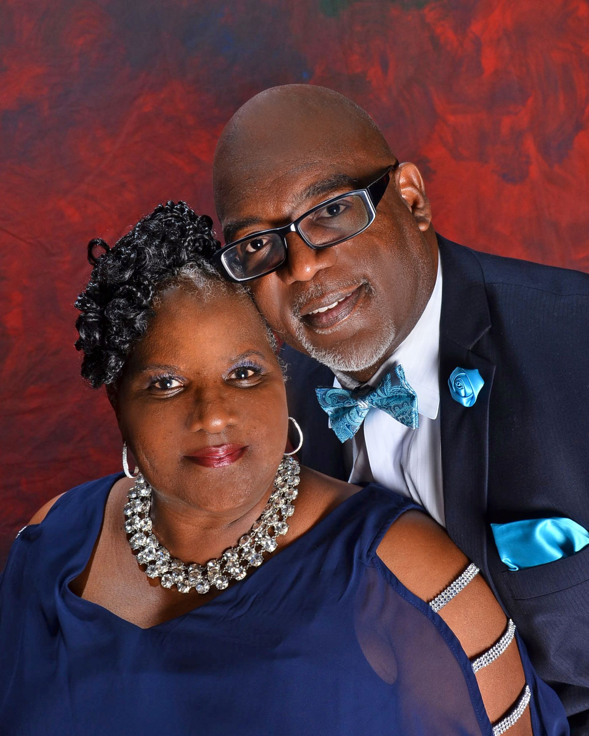 Couple in formal attire, smiling against a dark red background. The man wears glasses and a teal bow tie.