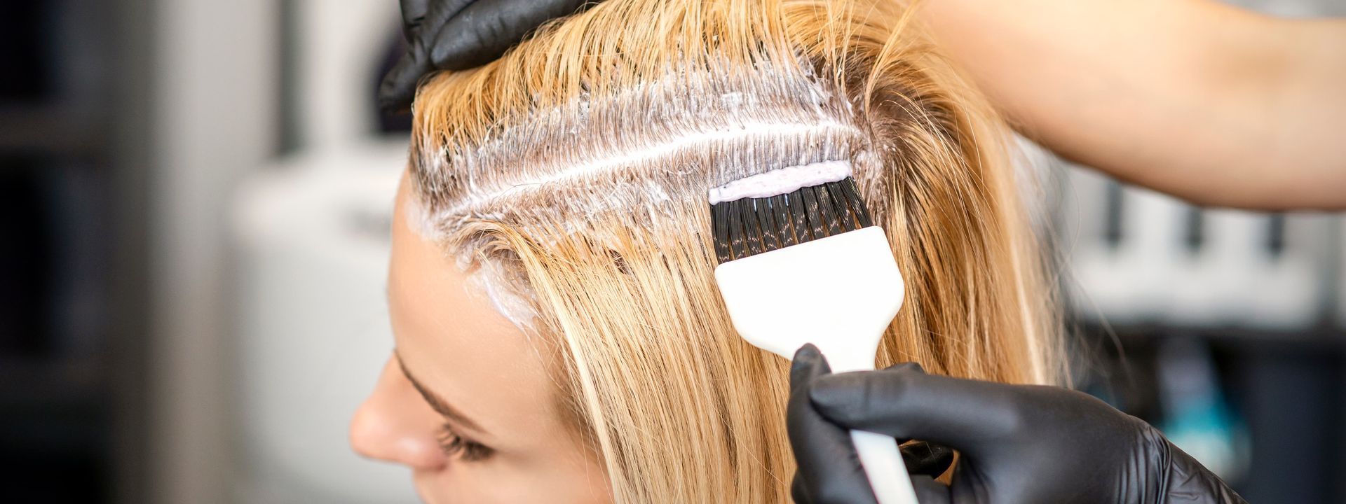 Hairdresser applying hair dye to a client's blonde hair using a brush, wearing black gloves.