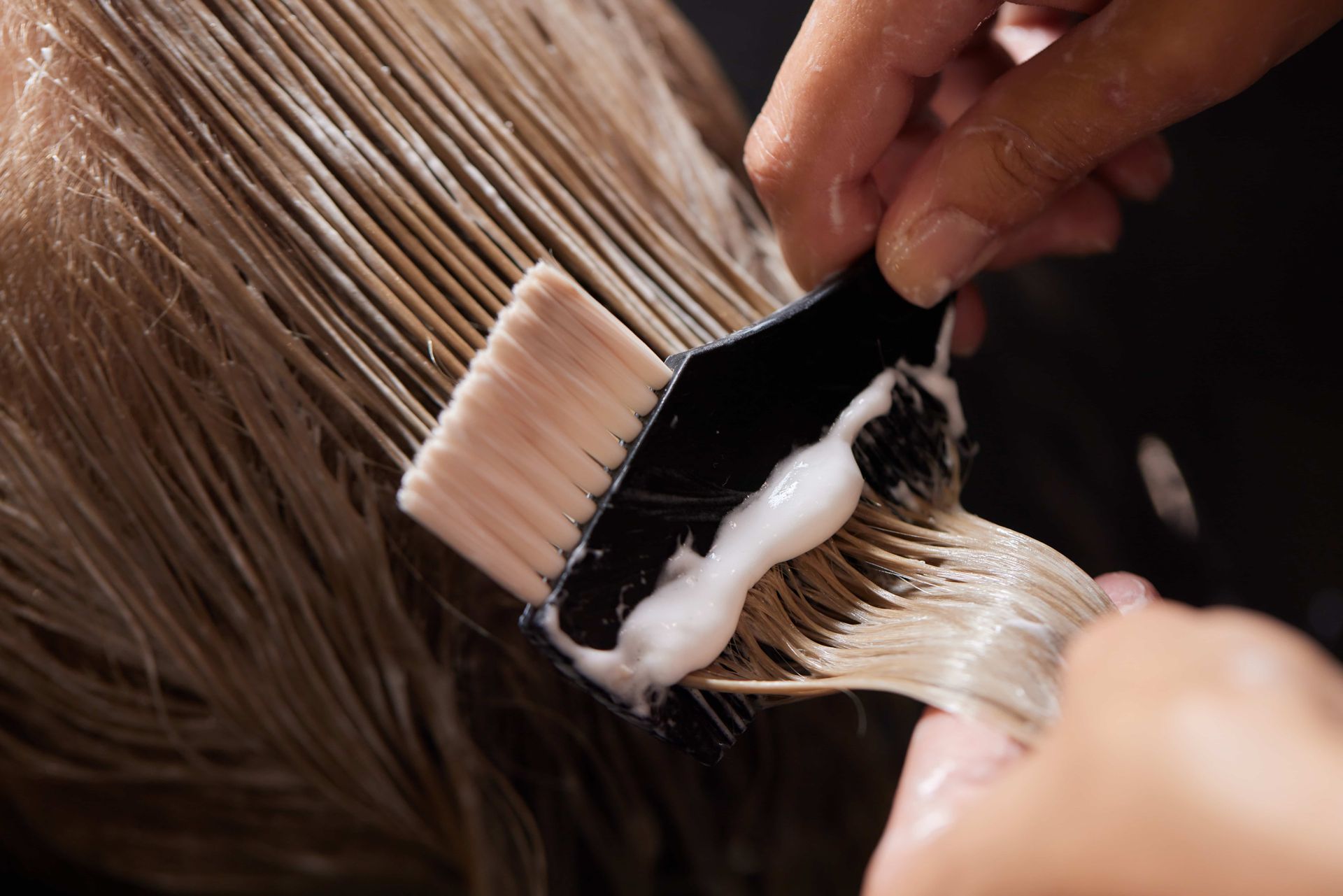 Hands applying hair dye with a brush to blonde hair.