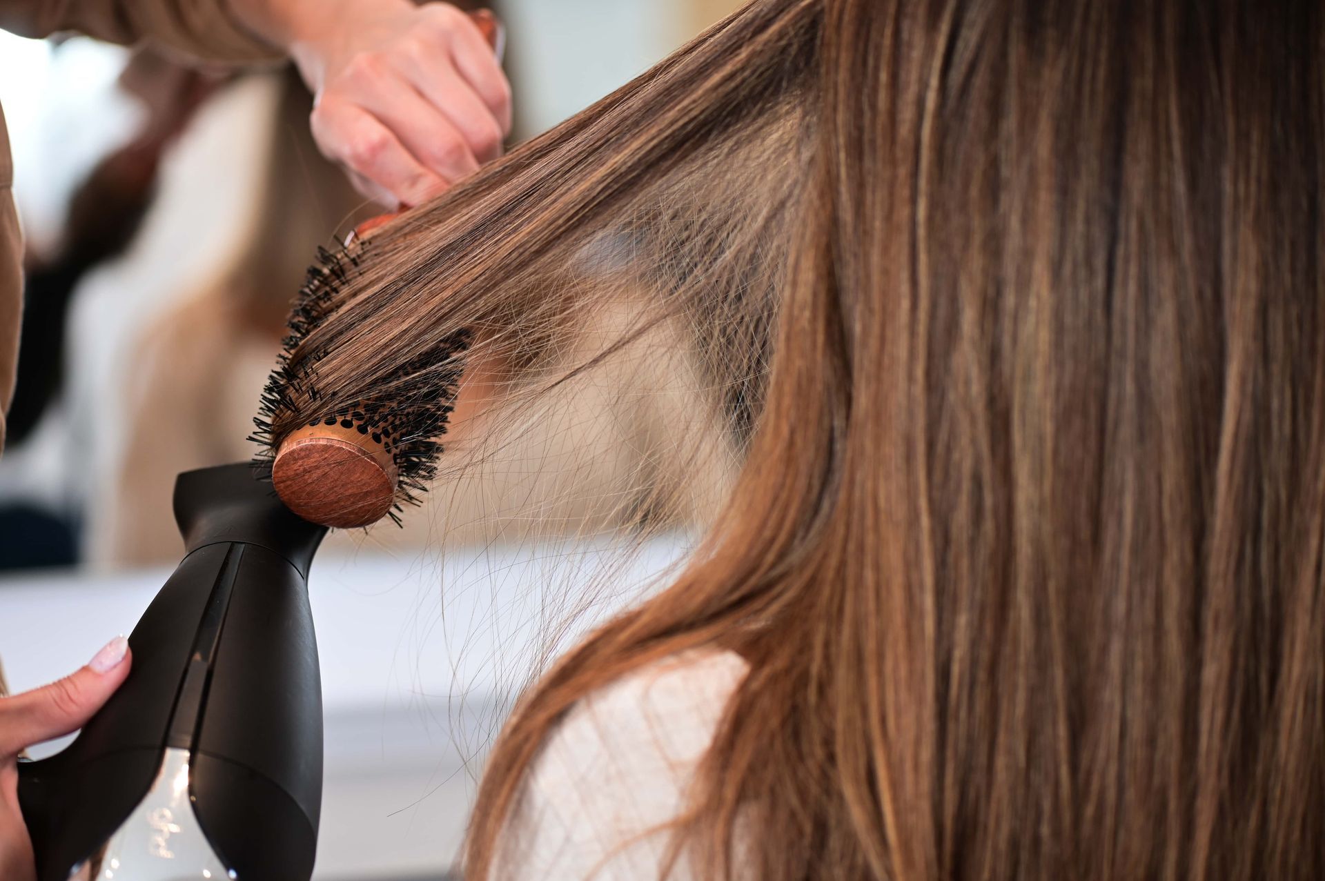 Hairdresser blow-drying a client's long, highlighted hair with a round brush in a salon.
