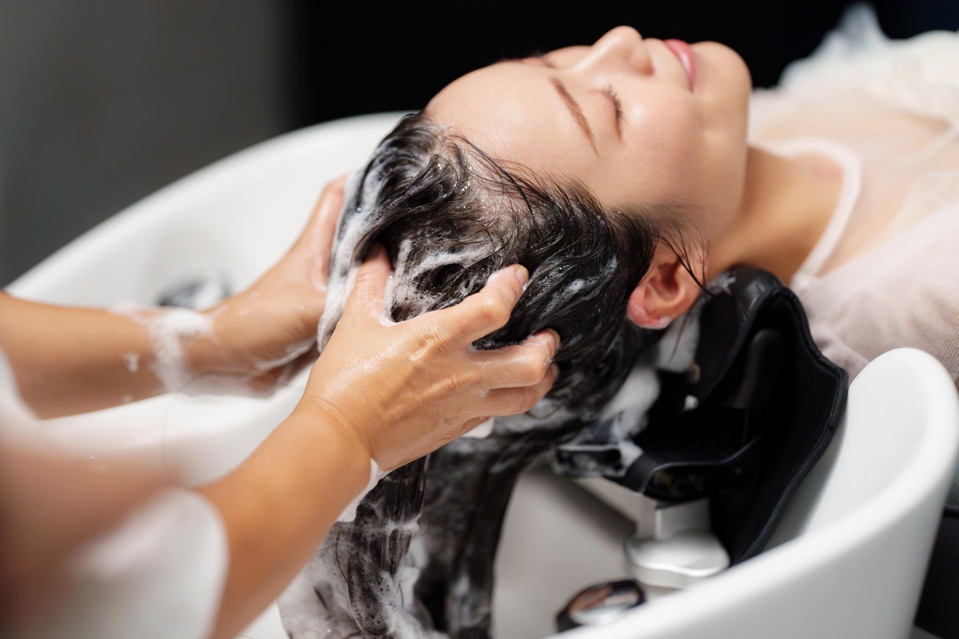 Woman getting hair washed at a salon, hands massaging shampoo into her dark hair over a white sink.