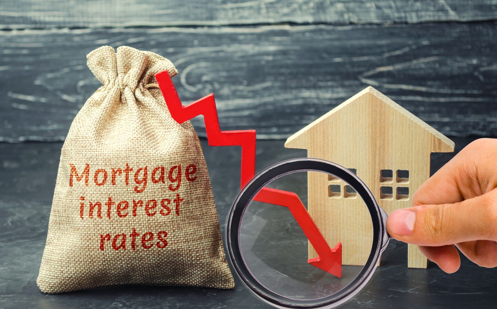 Hand holding magnifying glass over toy house and mortgage rates sack with red downward arrows