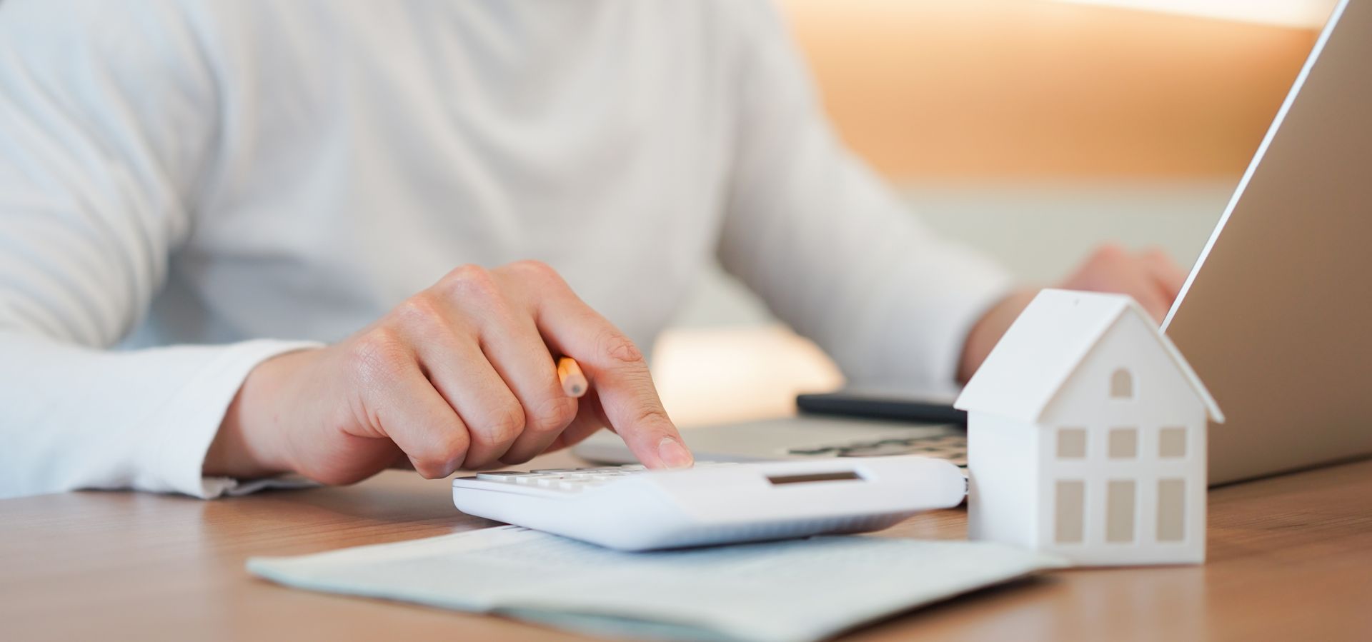 Person using a calculator beside a model house and laptop while reviewing home financing numbers.