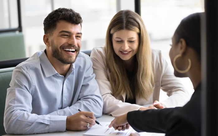 A couple smiling while signing mortgage documents with a loan consultant. A couple smiling while signing mortgage documents with a loan consultant.
