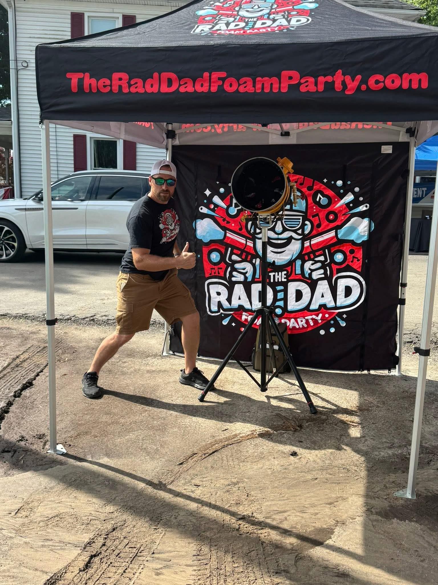 Man posing under a RAD DAD Foam Party tent beside a skull-themed banner outdoors