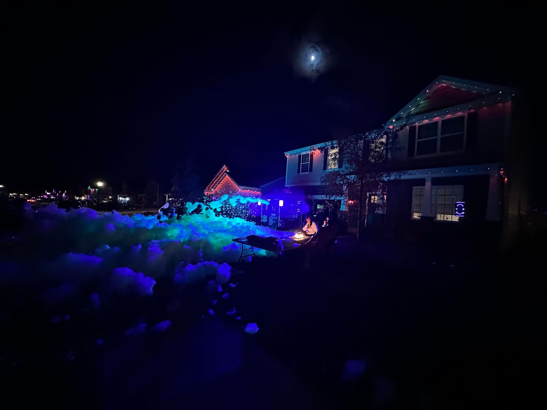 House and yard lit with blue and purple holiday lights at night under a bright moon