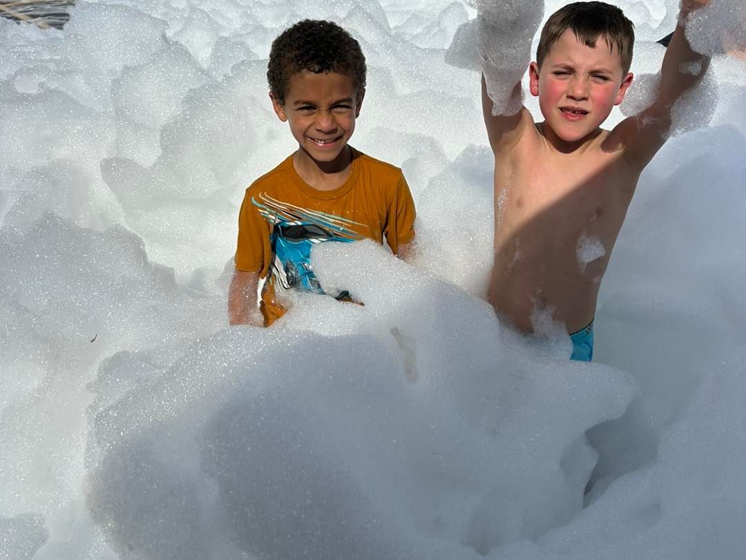 Two children playing in deep white foam or snow, smiling with arms raised outdoors.