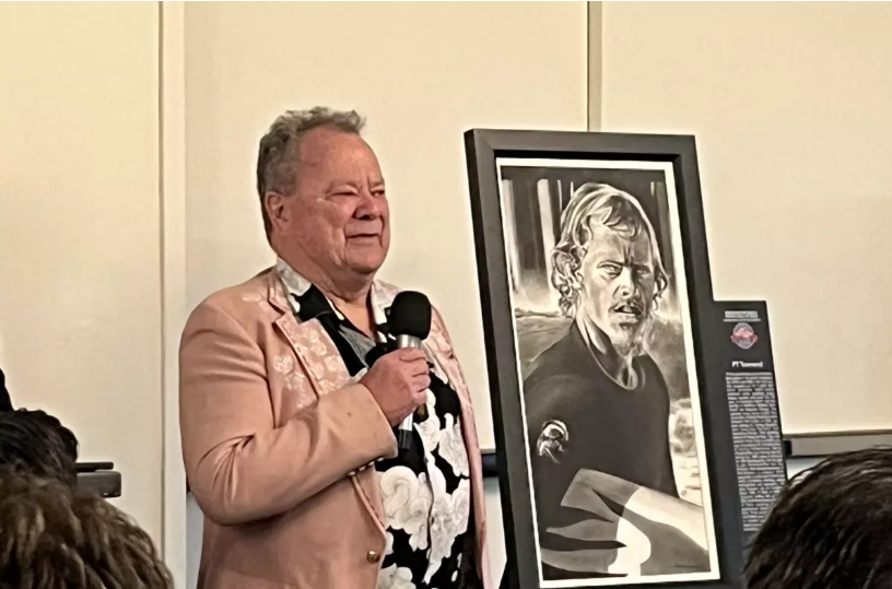 Daily Pilot article Man in pink jacket speaks into a microphone. Next to him is a framed black-and-white portrait.