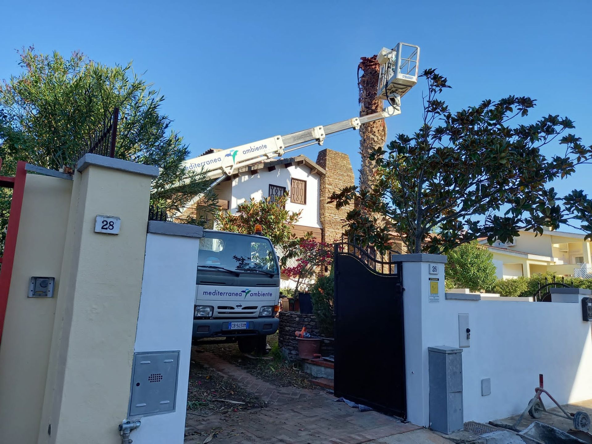 Un camion con un braccio sollevatore sta potando un'alta palma vicino a una casa con un cancello.