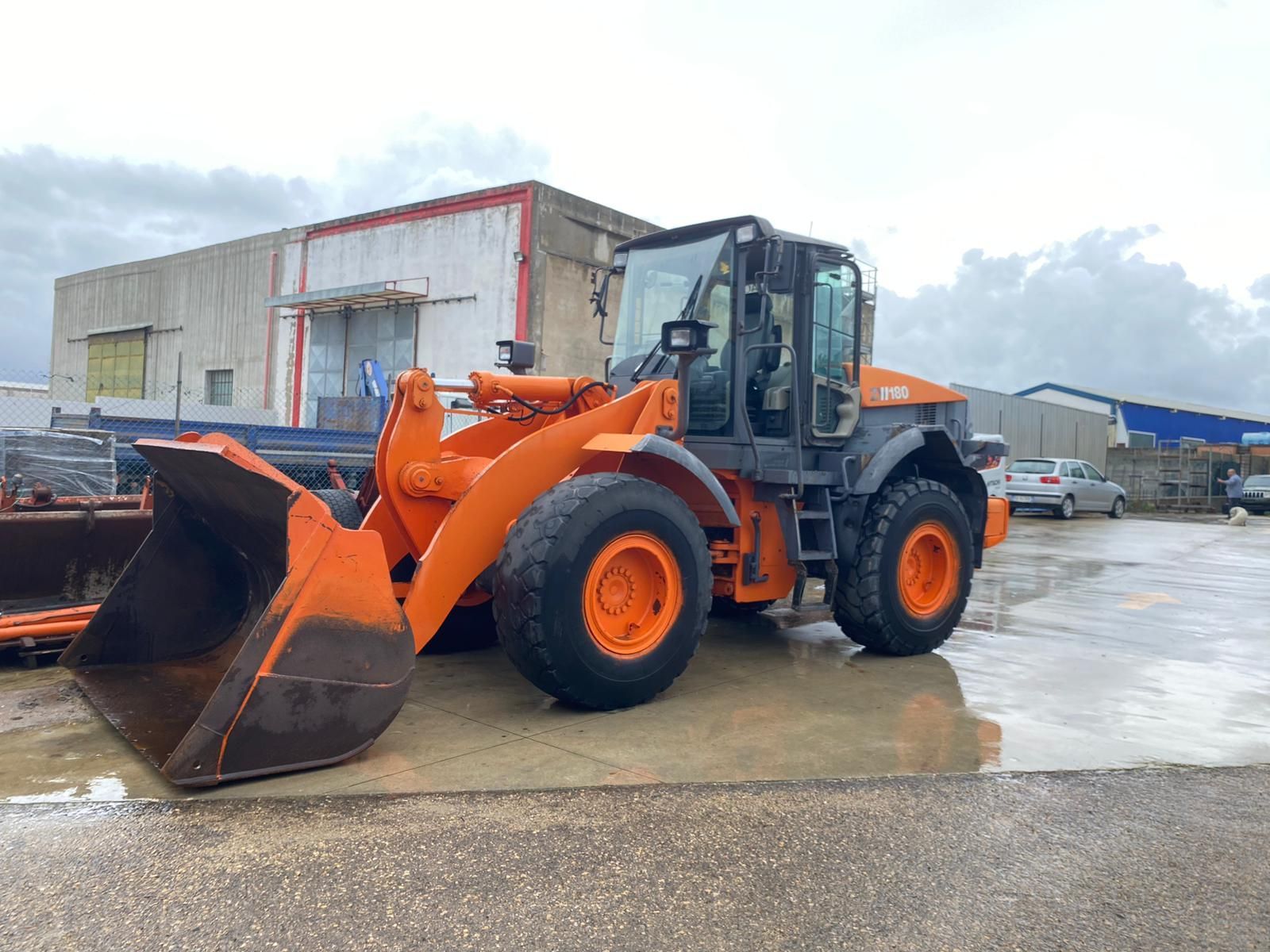 Pala gommata arancione Hitachi su una superficie pavimentata bagnata di fronte a un edificio sotto un cielo nuvoloso.