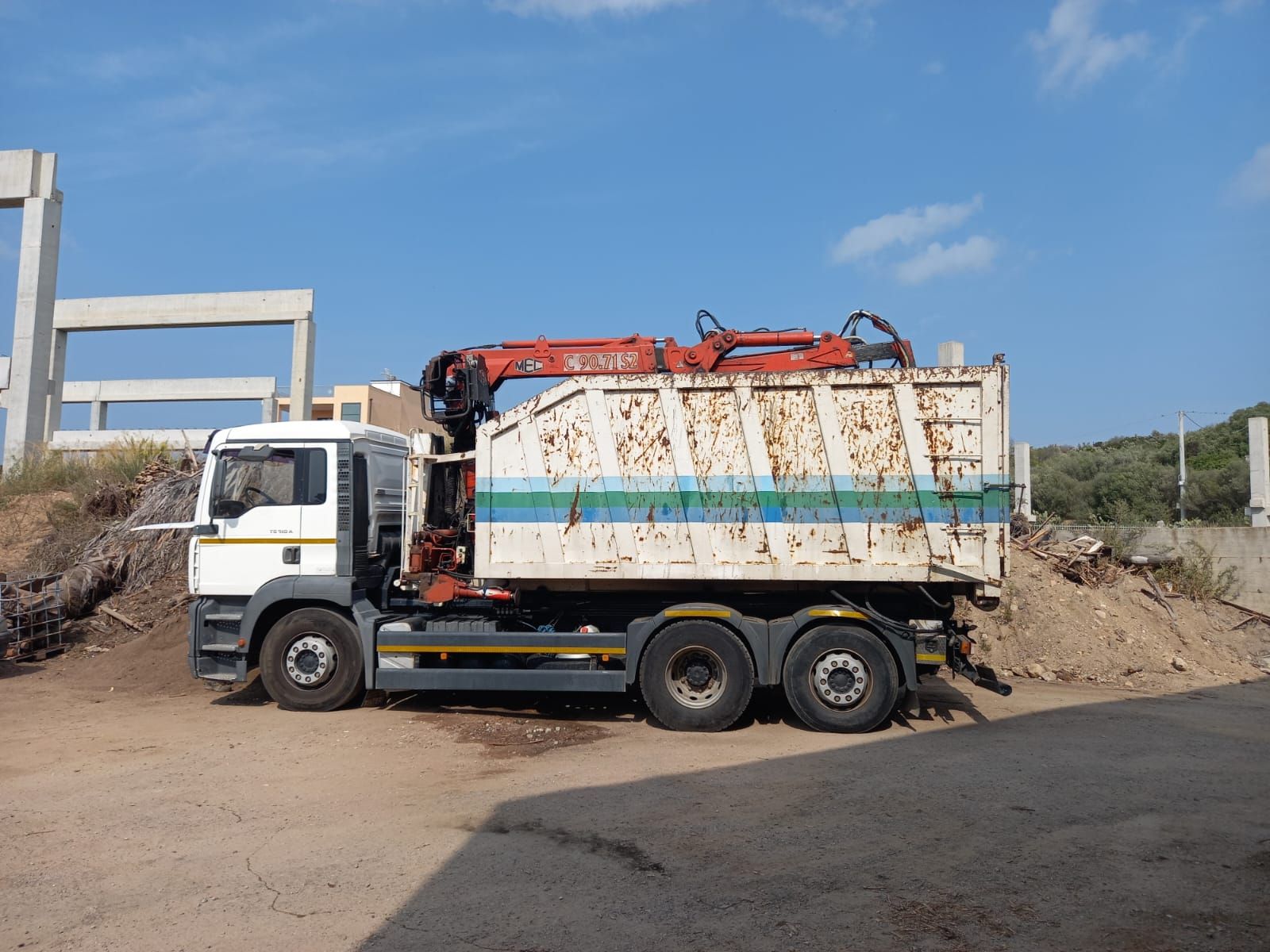 Un autocarro con cassone ribaltabile e una gru rossa parcheggiata accanto ai detriti edili sotto un cielo azzurro.