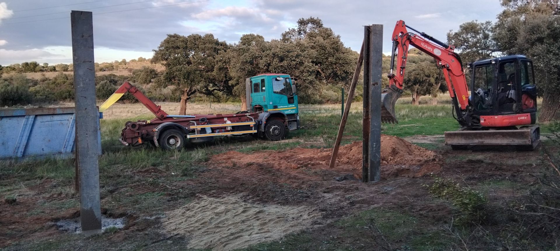 Cantiere: escavatore vicino ai pali, camion sullo sfondo, alberi verdi, cielo nuvoloso.