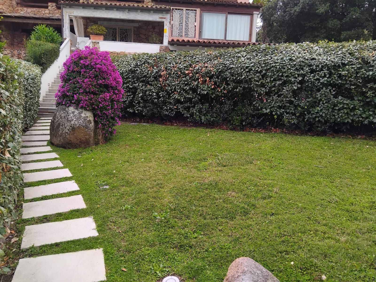 Un sentiero in pietra conduce a una casa con un rigoglioso prato verde, una siepe e un cespuglio fiorito.