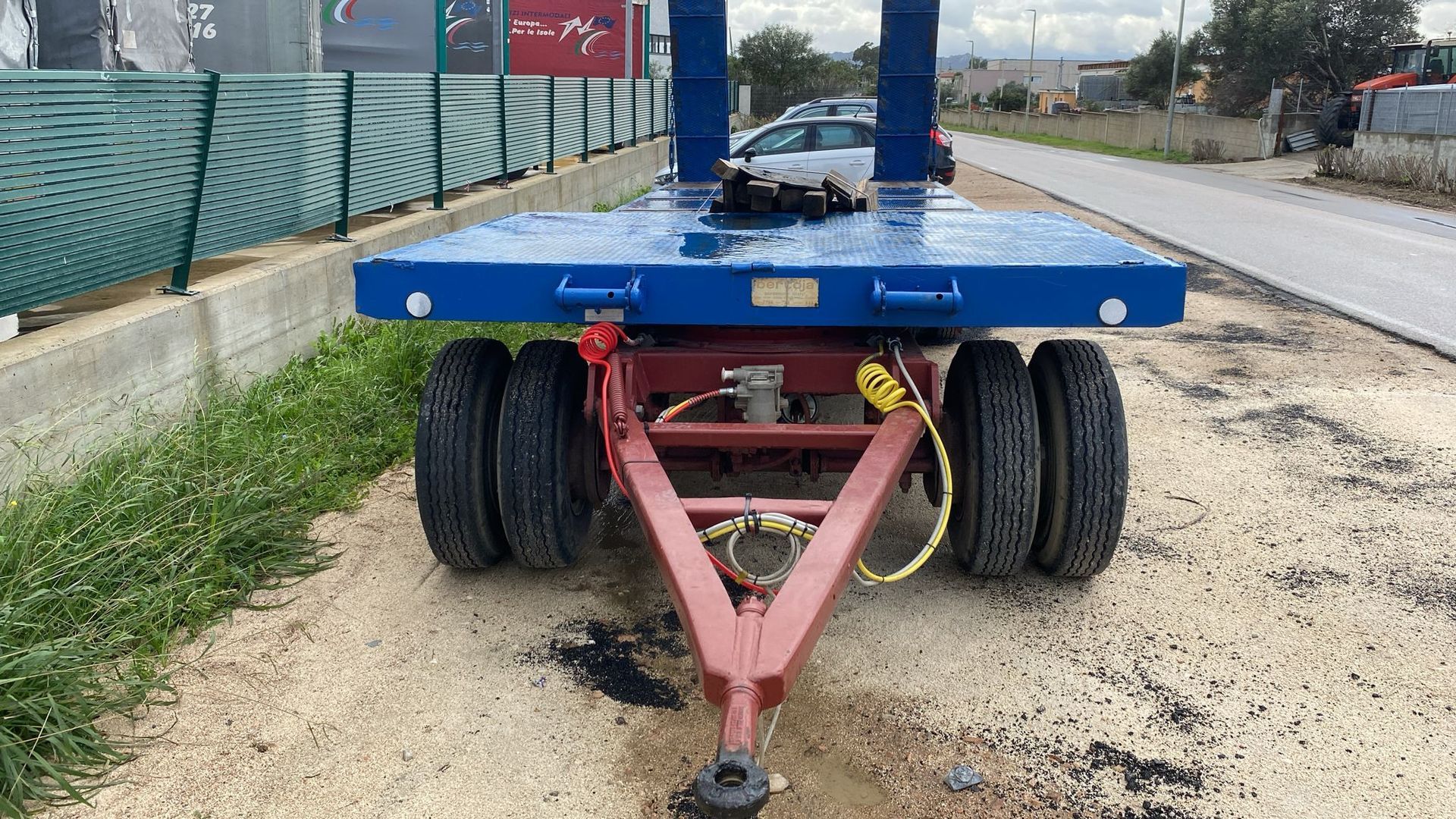 Rimorchio blu e rosso con pianale e sei pneumatici parcheggiato sulla ghiaia accanto a una strada.
