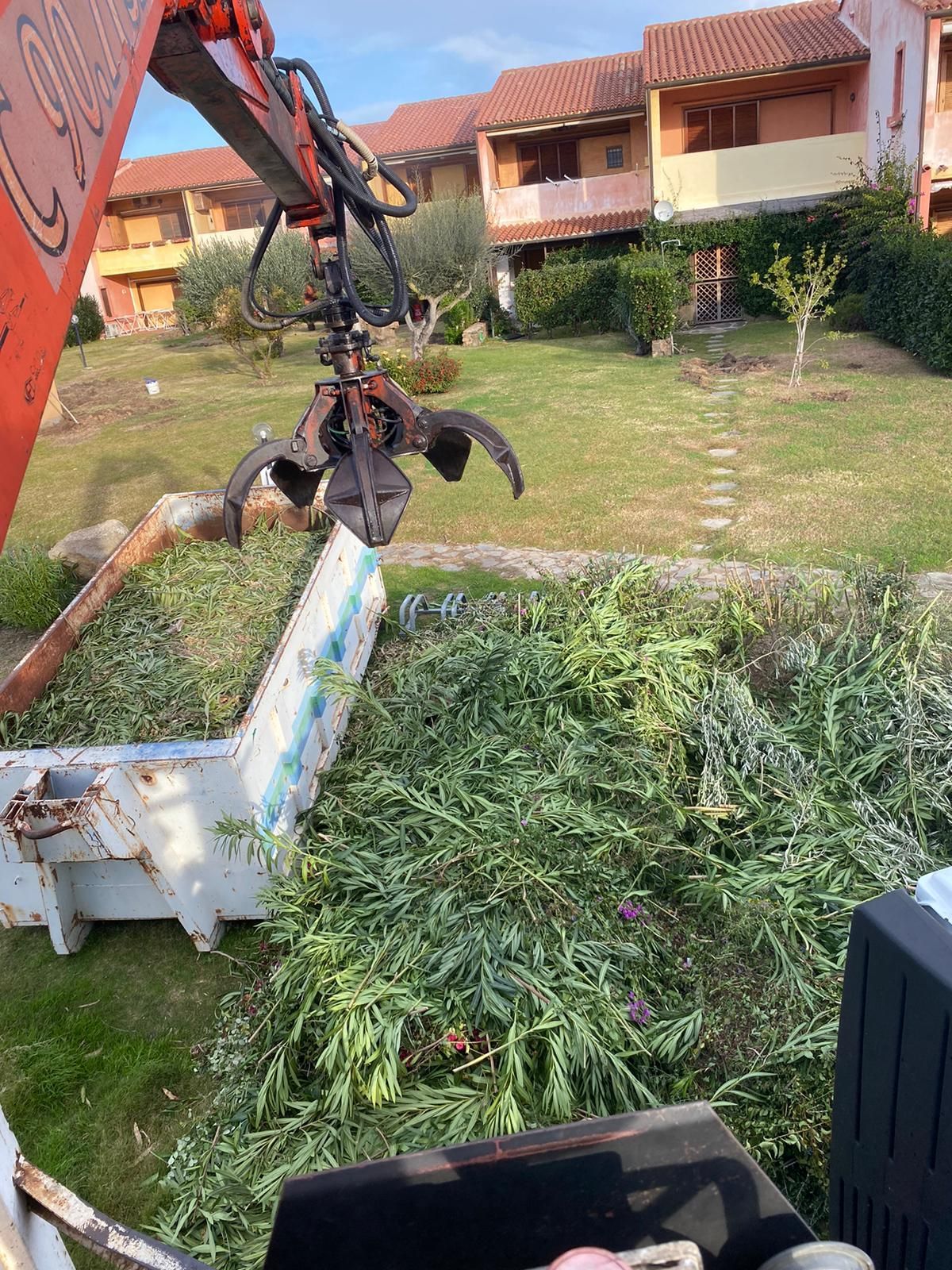 Una gru arancione che rimuove sterpaglie verdi da un contenitore bianco, vicino a case con prati verdi.