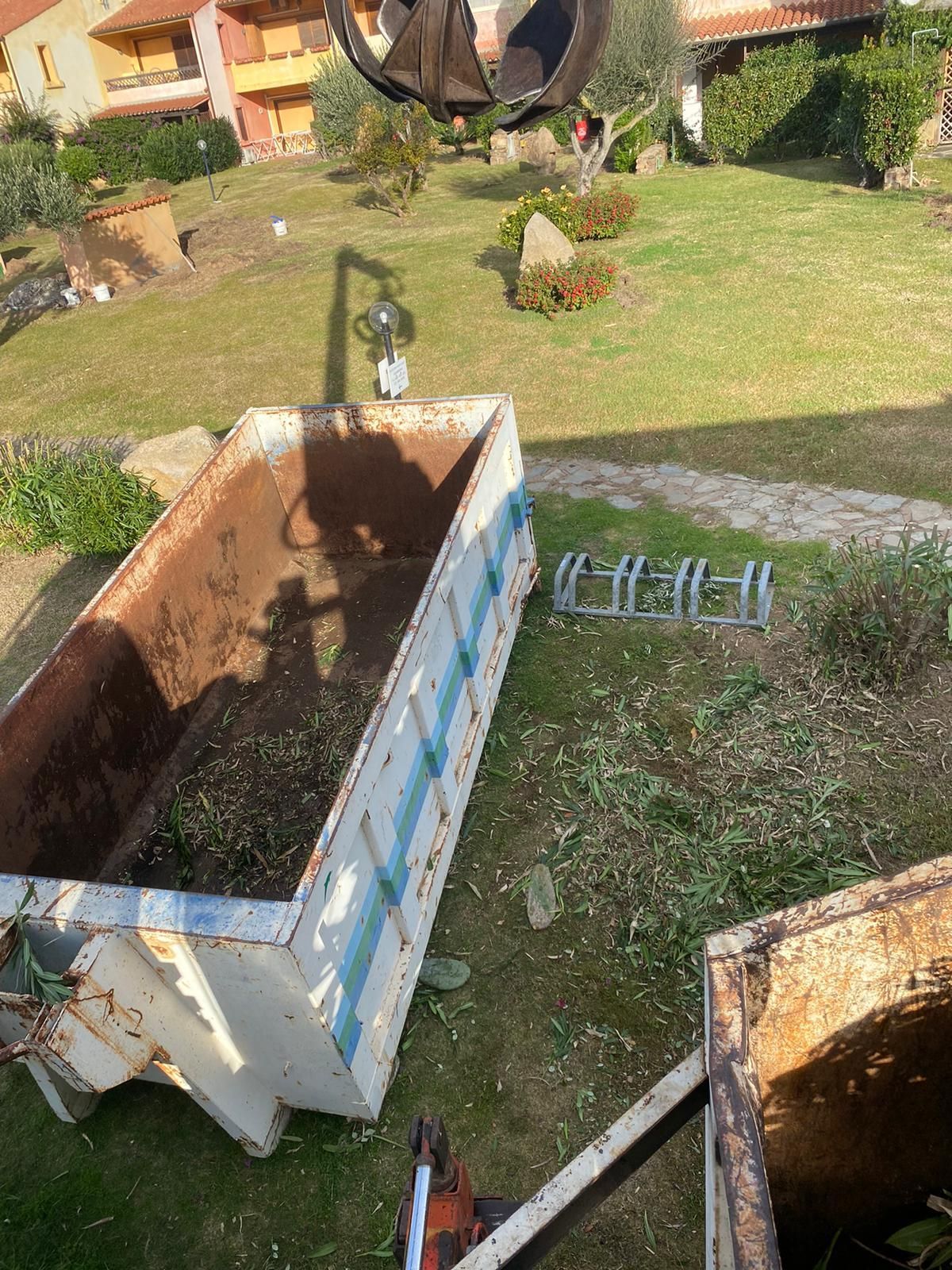 Un cassonetto arrugginito viene riempito da una pinza meccanica in un cortile erboso.