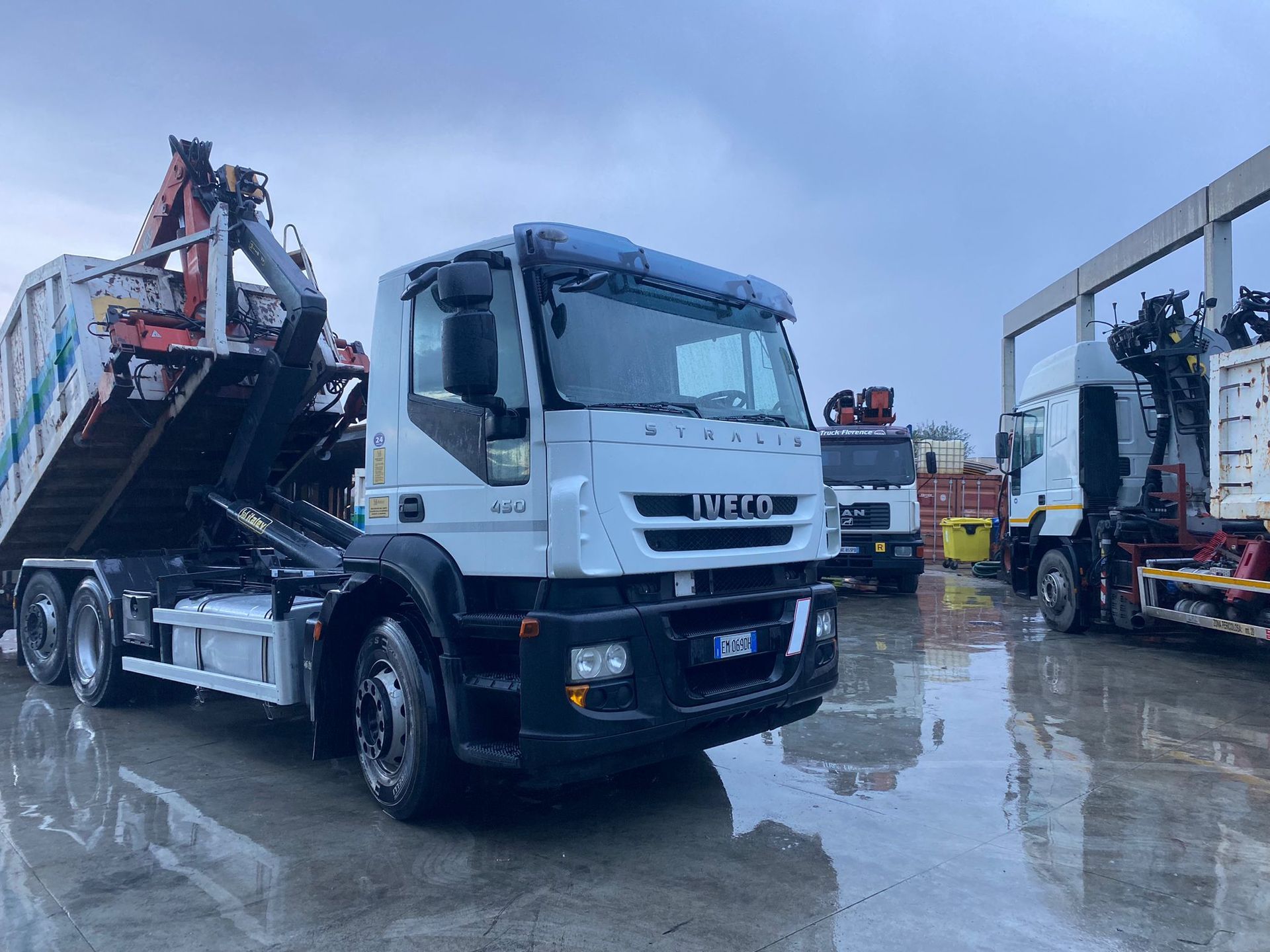 Camion della spazzatura Iveco bianco con cassone rialzato, altri camion in un cortile.