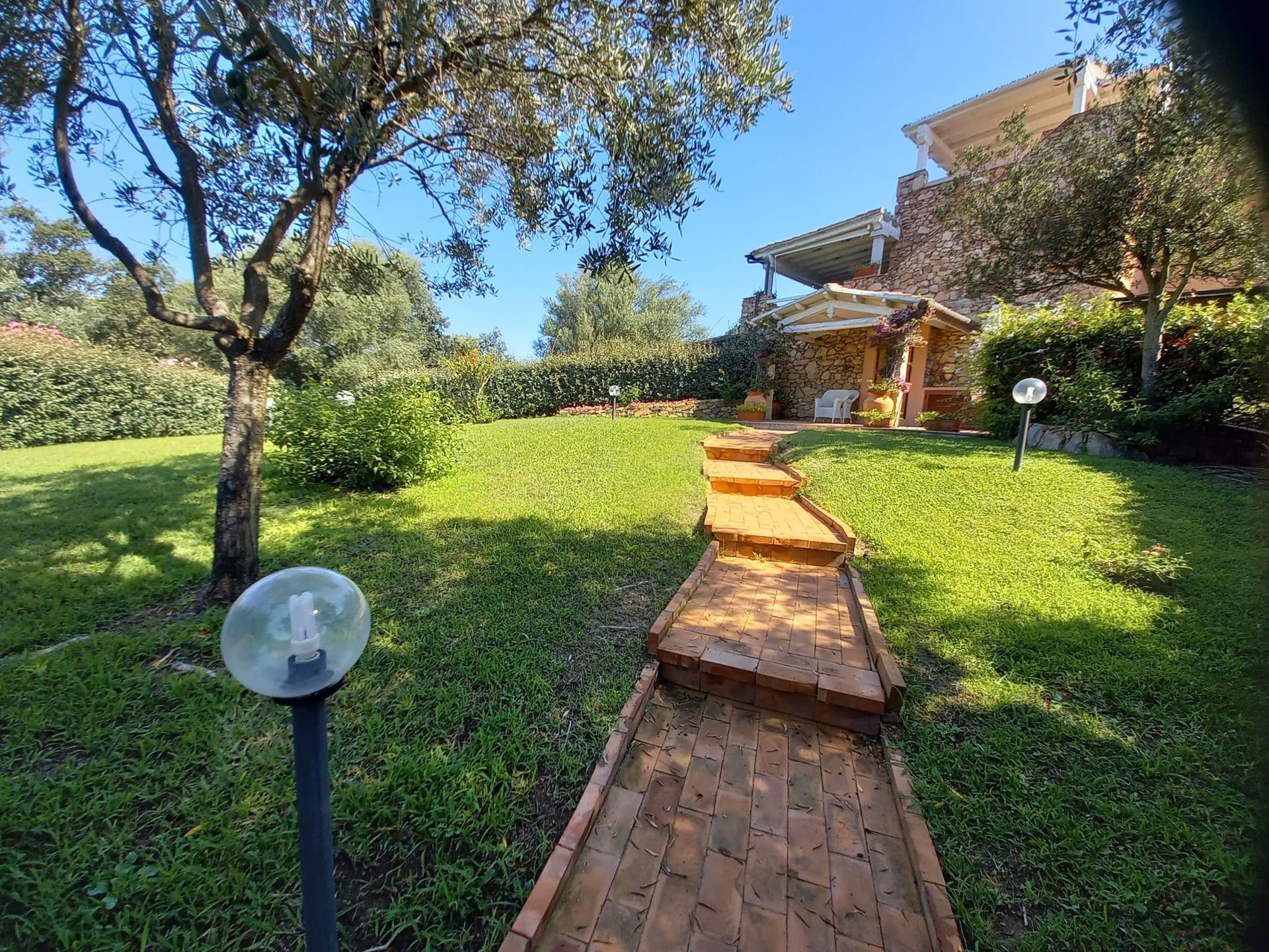 Sentiero in mattoni attraverso un prato verde verso un edificio in pietra con pergolato, giornata di sole.