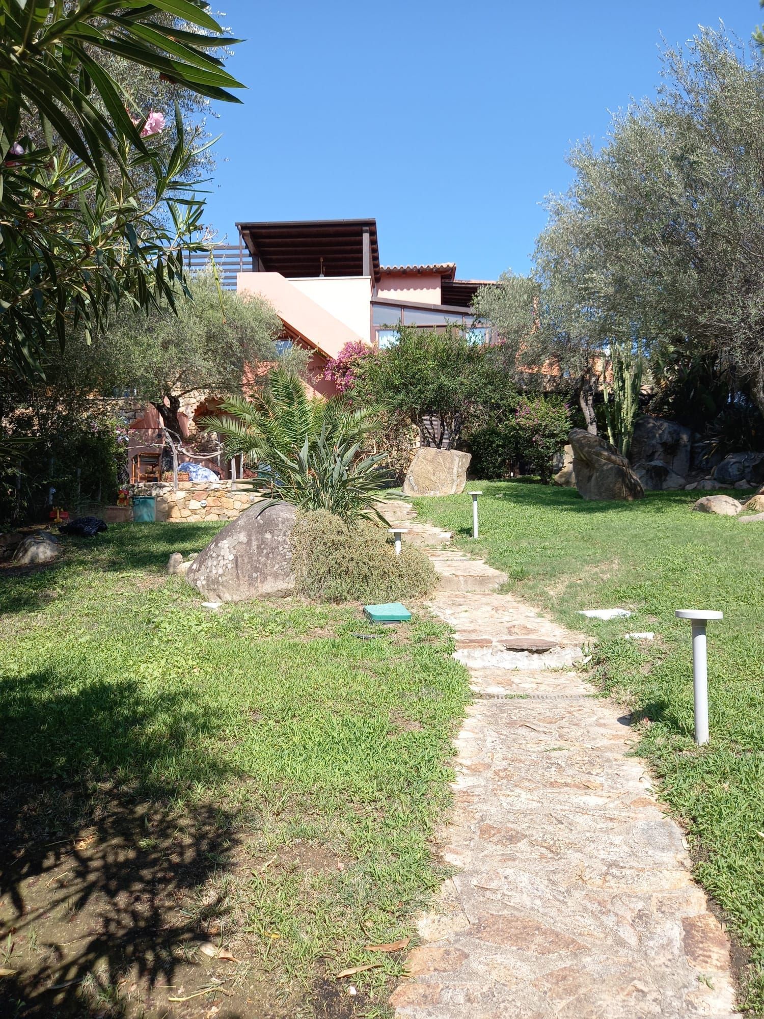 Sentiero in pietra attraverso un giardino verso un edificio di colore chiaro sotto un cielo azzurro.