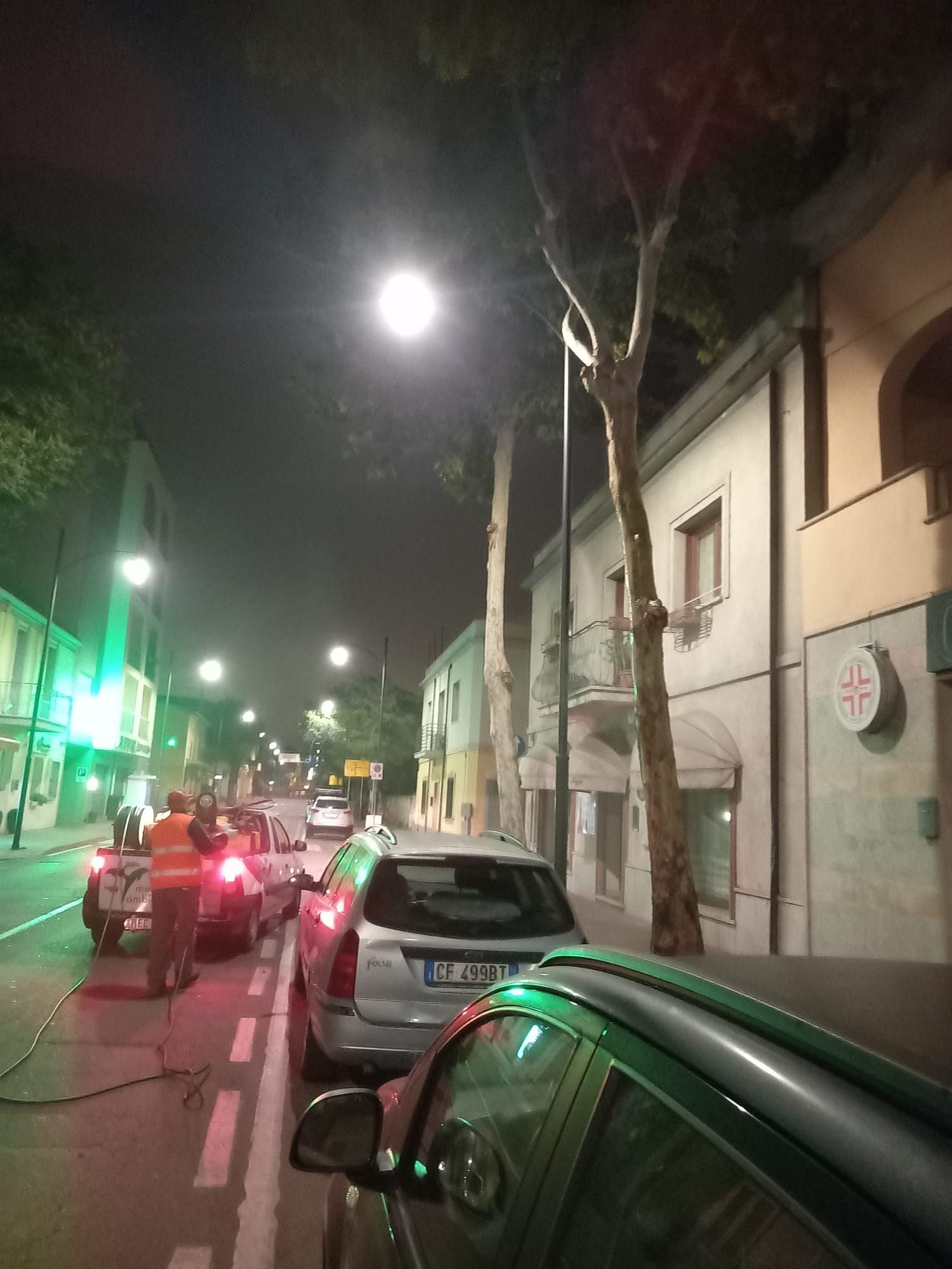 Scena di strada di notte con auto parcheggiate e operai vicino al semaforo verde. Edifici e alberi costeggiano la strada.