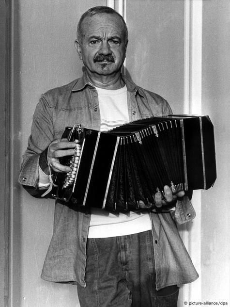 Hombre con bigote tocando un bandoneón, vistiendo una camisa vaquera y una camiseta blanca.