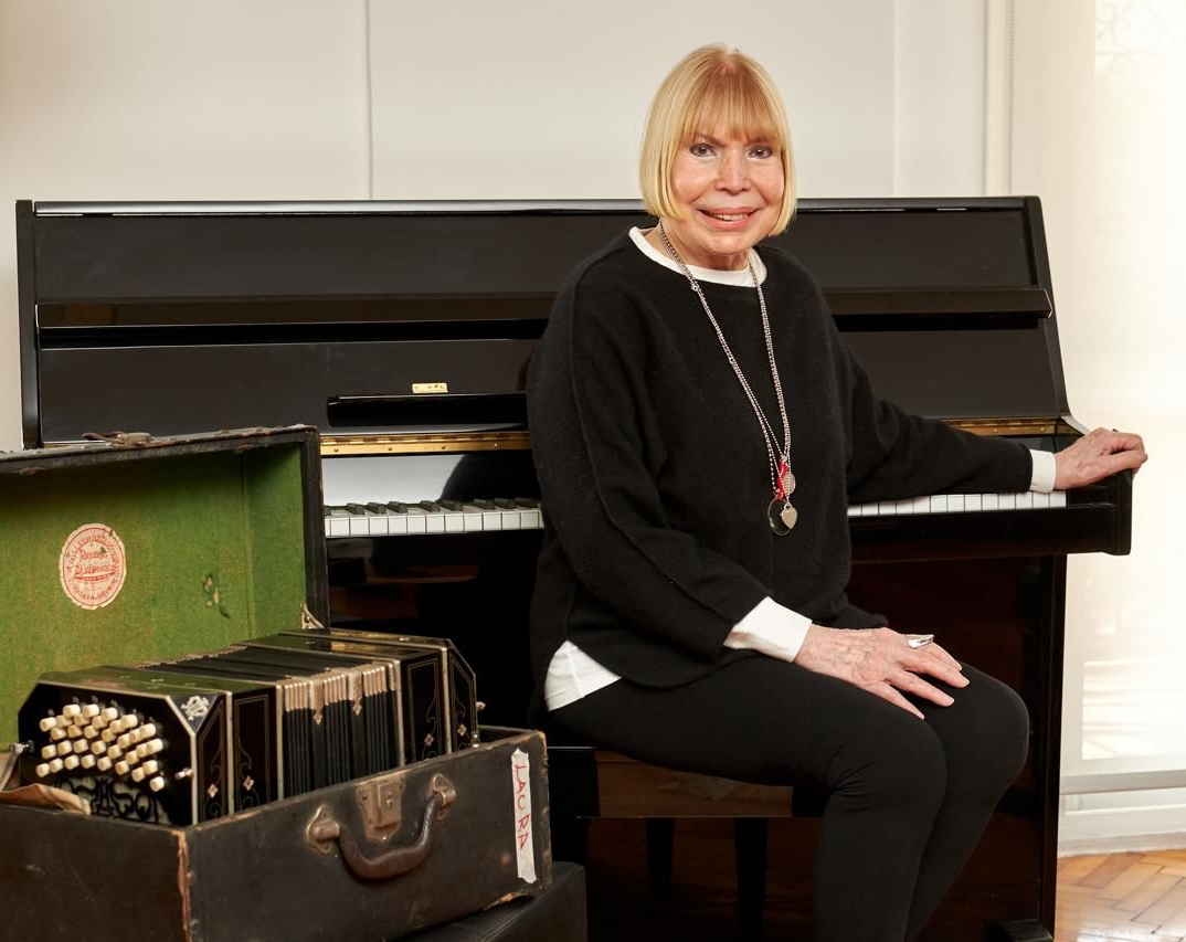 Mujer sentada al piano junto a un bandoneón. Vestida de negro, sonríe. Interior.