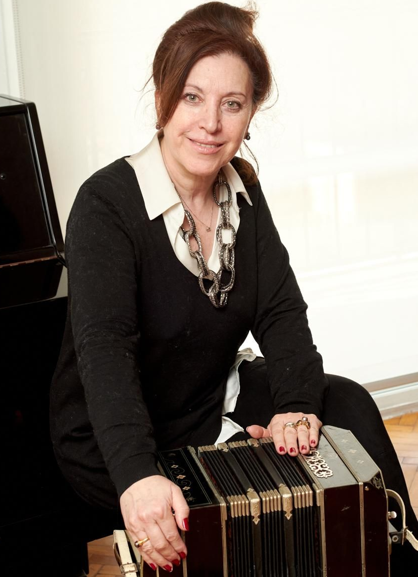 Mujer sosteniendo un bandoneón, sonriendo, frente a un piano, en el interior.