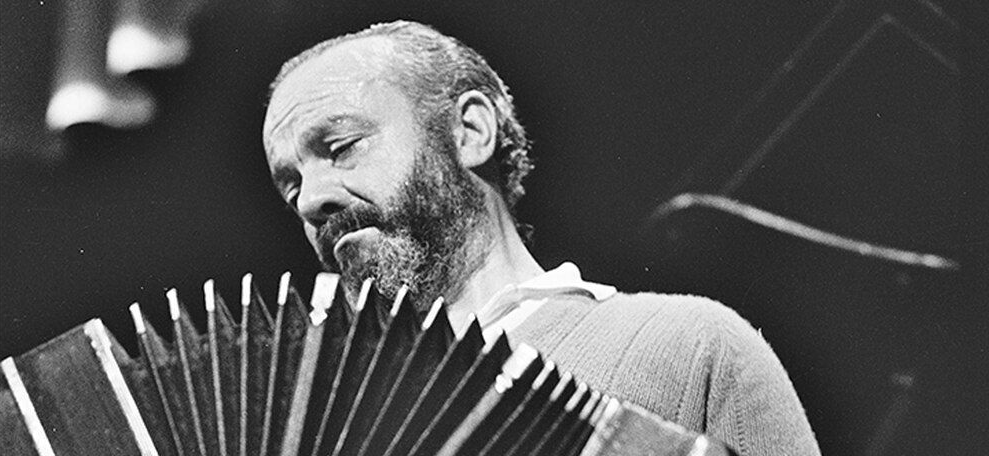 Un hombre calvo con barba tocando un acordeón delante de un fondo oscuro.
