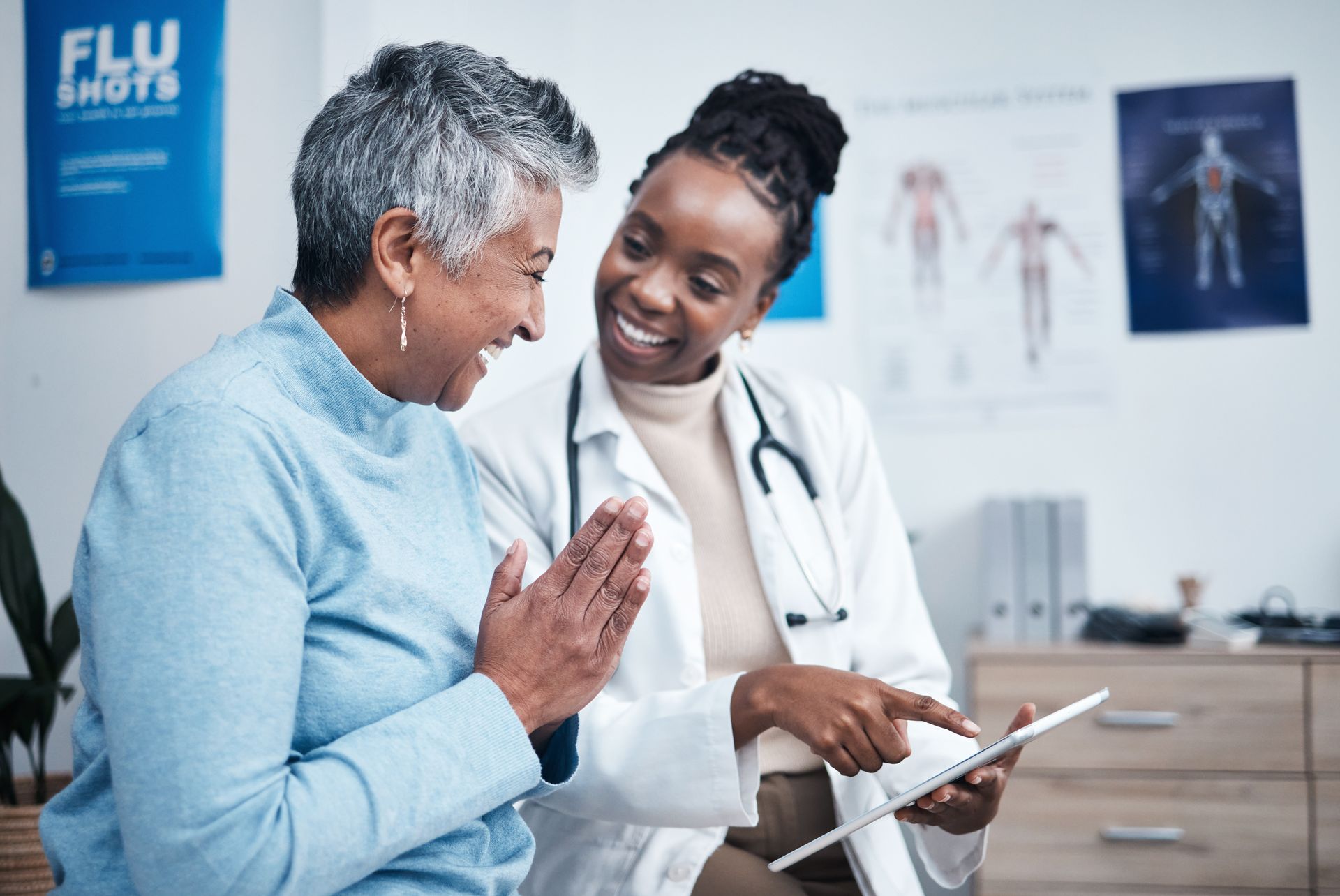 Doctor showing a tablet to a patient in a clinic, both smiling beside medical posters.