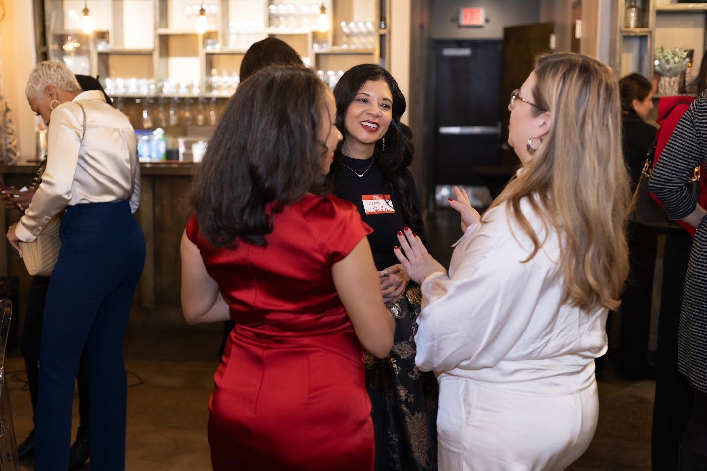 Three people chatting at a networking event in a warm-lit room, one in a red dress.