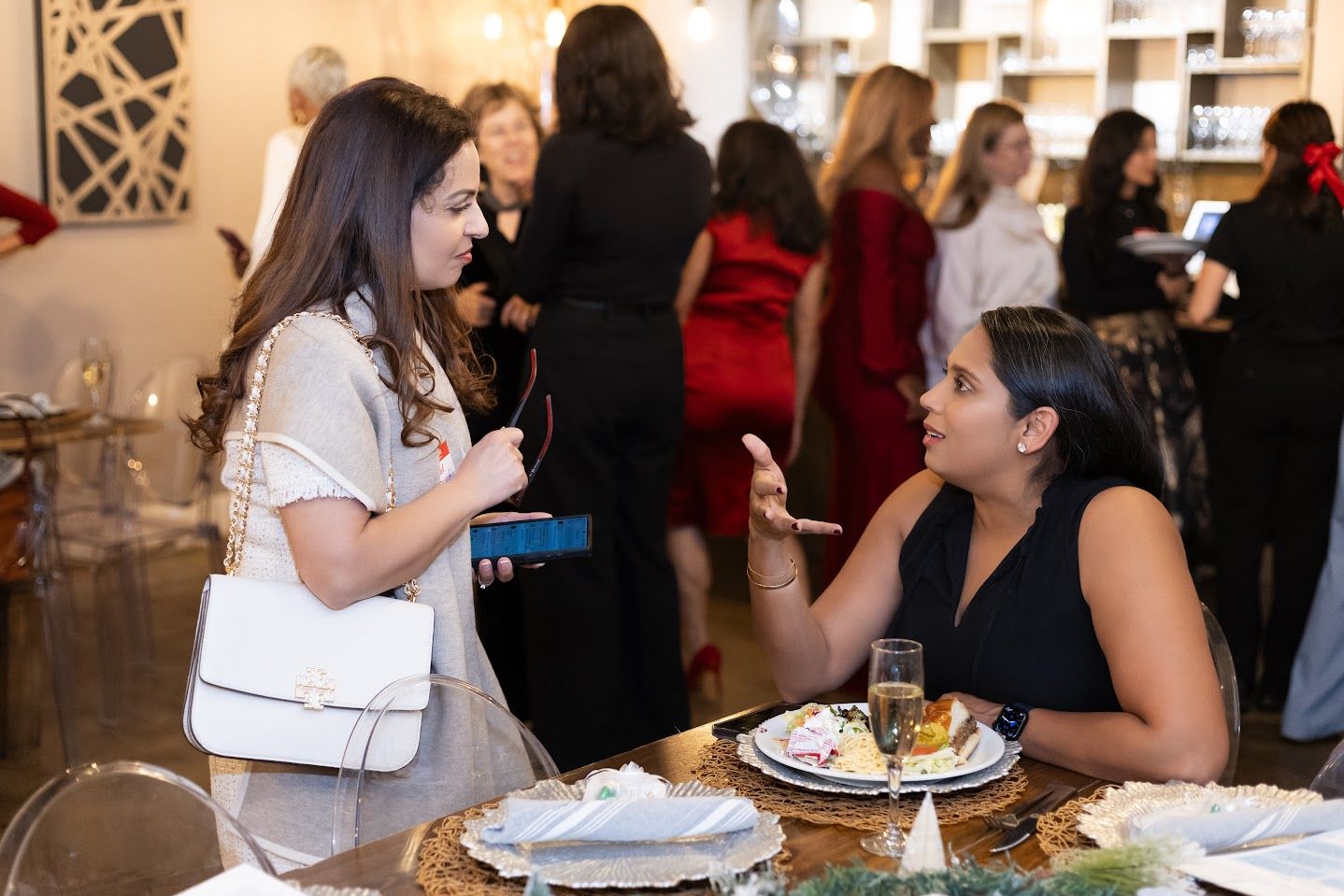 Two women chatting at a restaurant table with drinks and plates, while guests mingle in the background