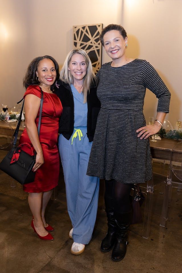 Three women posing indoors at a festive event, with holiday decor and smiling at the camera.