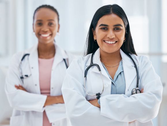 Two smiling doctors in white coats with stethoscopes, standing with arms crossed in a bright clinic