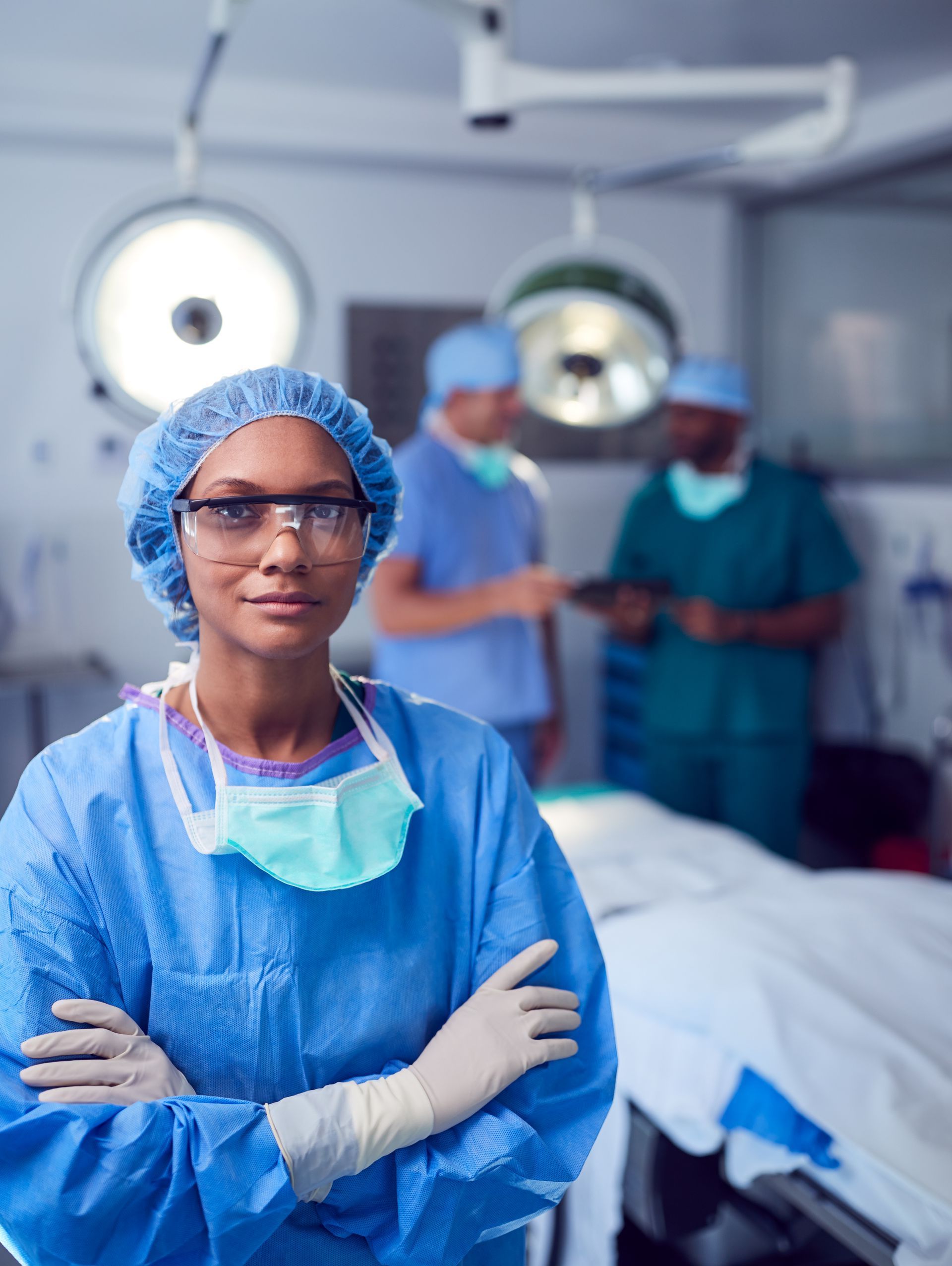Healthcare worker in blue protective gear stands in an operating room with surgical staff in the background
