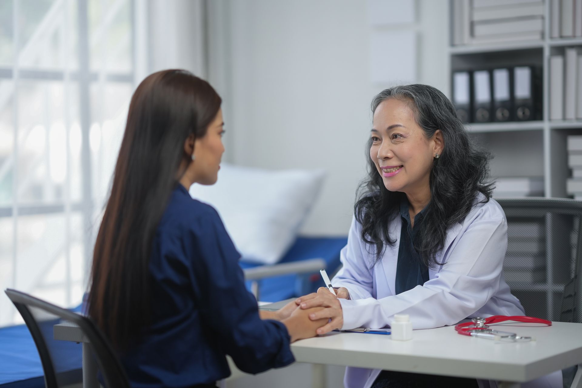 Two women talk at a desk in a bright office, one smiling and holding the other’s hands.