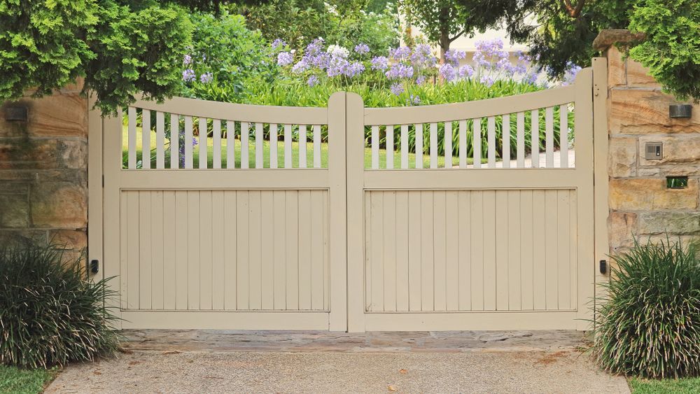 Cream-colored wooden gate with vertical slats and arch-shaped top, set in a stone wall, opening onto a garden path — Sunshine Gates in Noosa, QLD