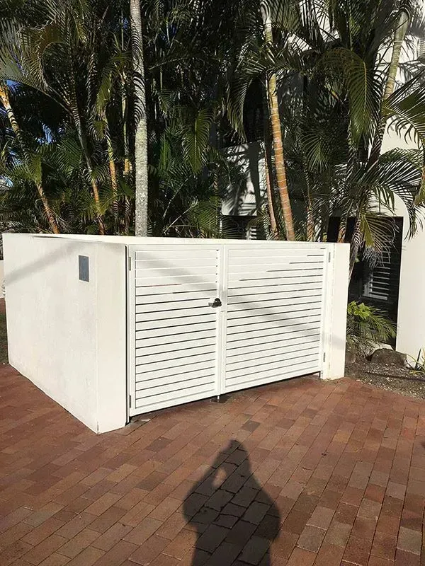 A white fence is sitting on top of a brick driveway next to a palm tree. — Sunshine Gates in Kunda Park, QLD