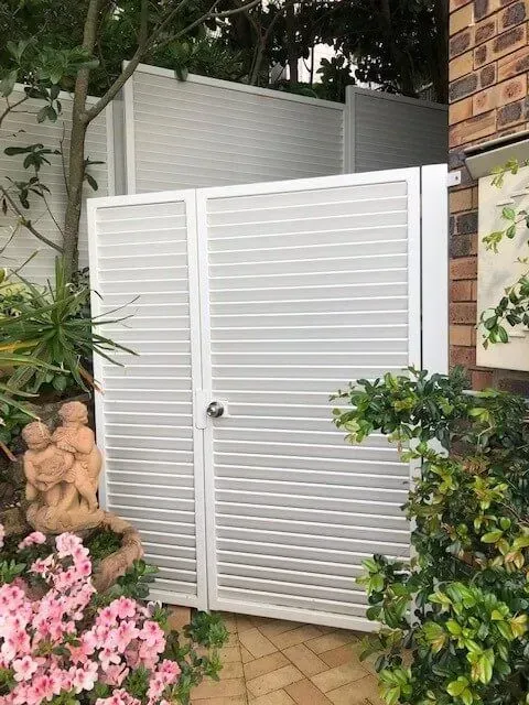 A white gate is sitting next to a brick building in a garden. — Sunshine Gates in Kunda Park, QLD
