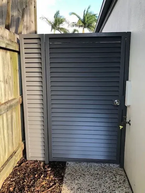A black gate is sitting next to a wooden fence.— Sunshine Gates in Kunda Park, QLD