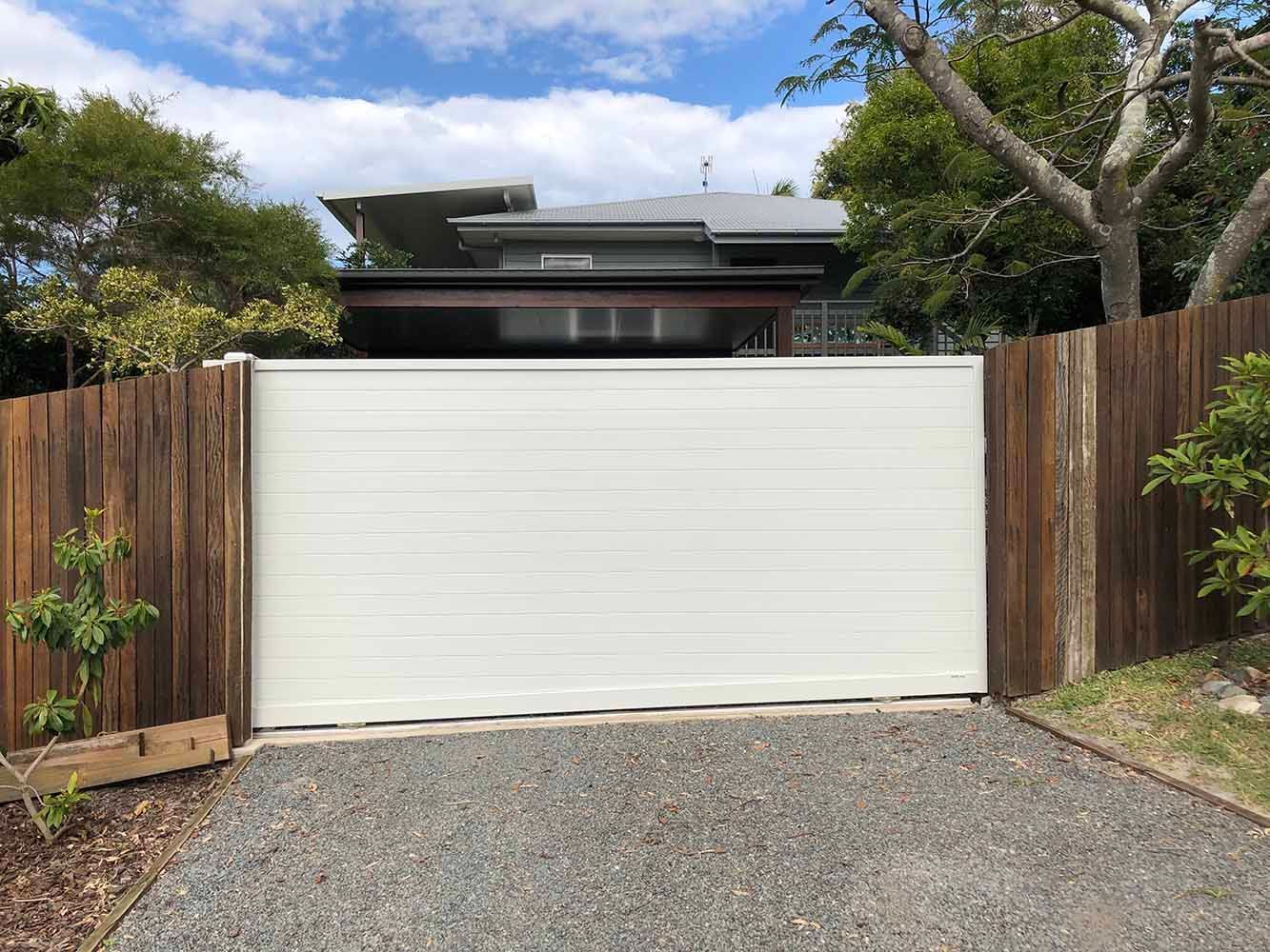 A white sliding gate is leading to a house behind a wooden fence. — Sunshine Gates in Kunda Park, QLD