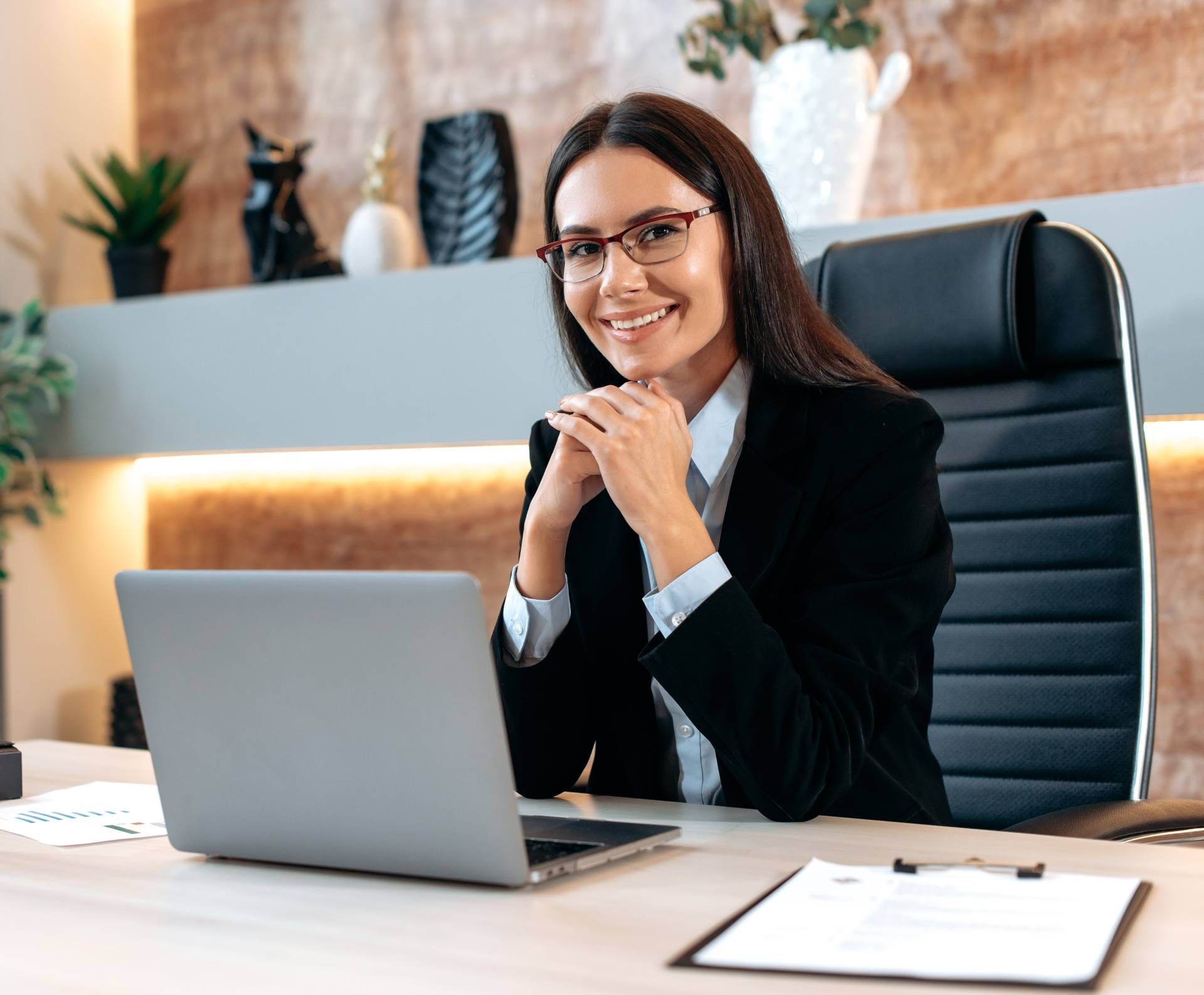 Woman in business attire smiles at desk with laptop, office setting.