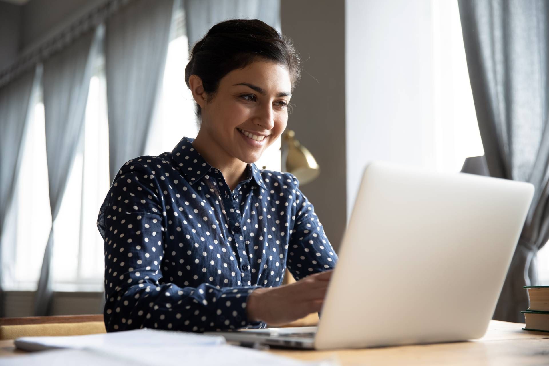 Woman smiling, working on a laptop at a desk near a window.