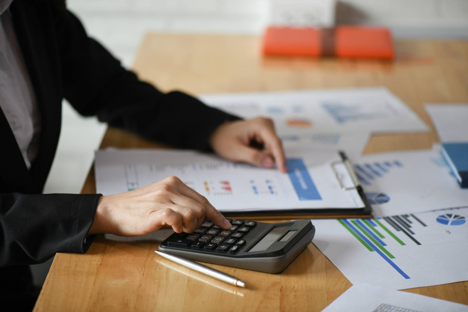 Person in a suit calculating with a calculator, reviewing papers with charts and graphs on a wooden desk.